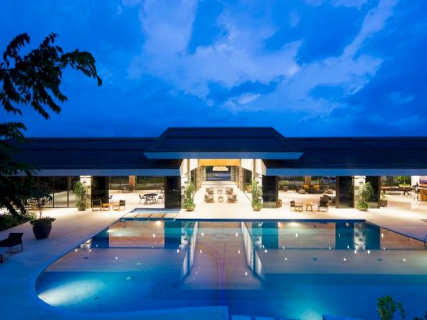A beautifully lit large outdoor pool area at a modern resort with a central building, surrounded by lounge chairs and scenic nighttime sky.