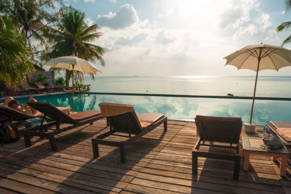 A serene poolside scene with lounge chairs, umbrellas, tropical trees, and a view of the ocean under a partly cloudy sky, exuding tranquility.