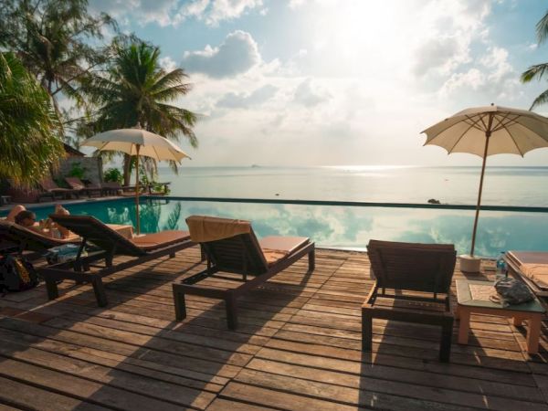 A serene poolside scene with lounge chairs, umbrellas, tropical trees, and a view of the ocean under a partly cloudy sky, exuding tranquility.