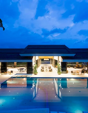 This image shows a luxury resort or hotel with a large, illuminated pool and modern architecture, set against a twilight sky.
