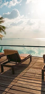 Two sun loungers and umbrellas on a wooden deck overlooking a tranquil pool with a scenic ocean view, surrounded by palm trees under a partly cloudy sky.