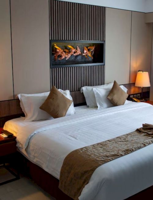 A well-decorated hotel room features a large double bed flanked by bedside tables with lamps, a wall-mounted fireplace, and a large window with curtains.