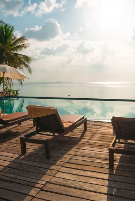 Poolside deck with lounge chairs, umbrellas, and a scenic view of a calm sea under a bright sky. Tropical setting with palm trees.