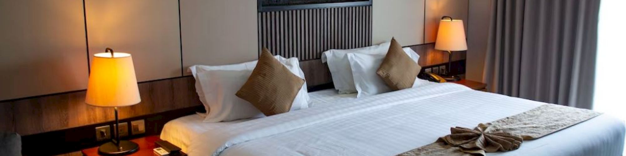 The image shows a well-decorated hotel room with a large bed, bedside tables with lamps, and a window with curtains.
