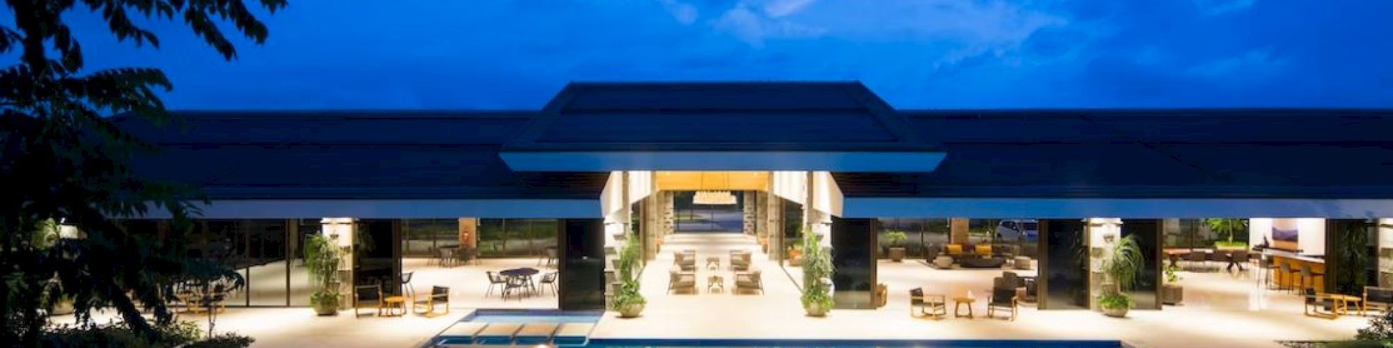 A luxurious building with a large swimming pool and outdoor seating, illuminated at night under a beautiful blue sky.