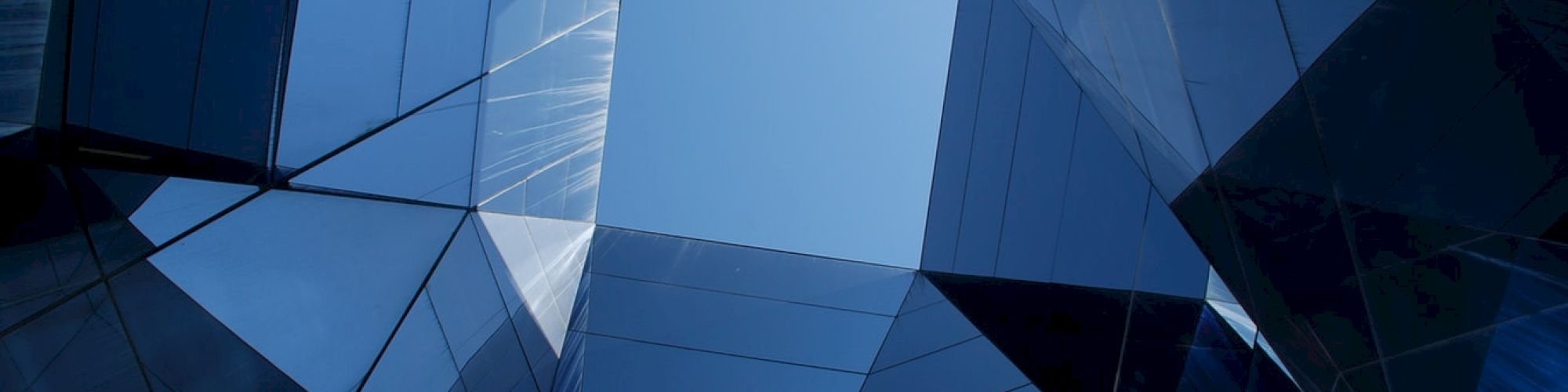 This image features a geometric, abstract view looking up through tall, reflective glass structures towards a bright blue sky.