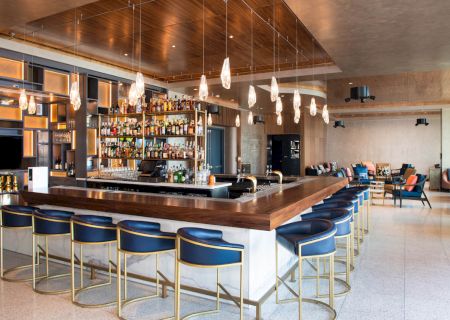 A modern bar with blue upholstered stools, a well-stocked liquor shelf, and pendant lighting. Lounge area with seating in the background.