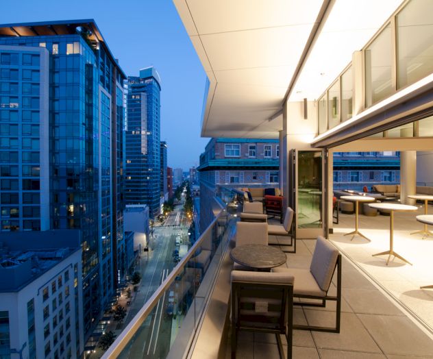 A modern outdoor terrace with seating and tables, overlooking a city street with tall buildings at dusk. The area is well-lit and inviting.