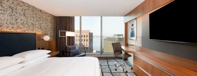 A modern hotel room features a large bed, wall-mounted TV, desk, and window with a cityscape view.