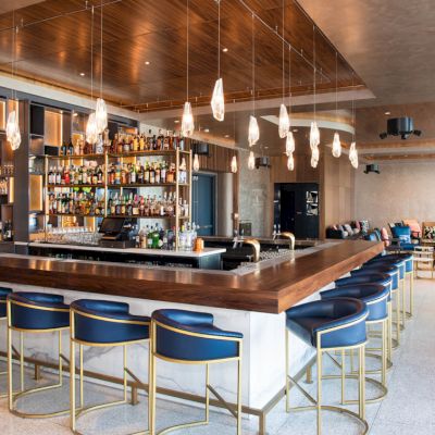 A modern bar area with a wooden counter, blue and gold bar stools, hanging lights, and shelves stocked with bottles. Comfortable seating is visible in the background.