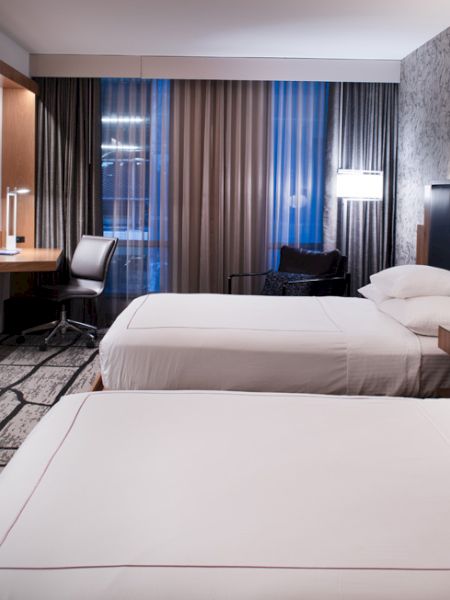 A modern hotel room with two beds, a large TV, a desk, and a chair. The decor is stylish, featuring contemporary furniture and soft lighting.