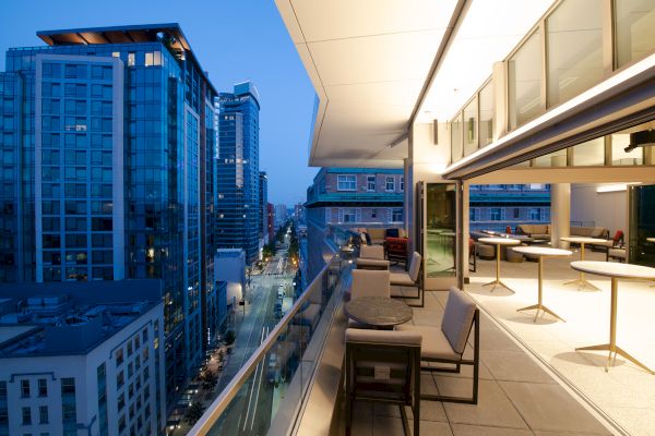 The image showcases a modern cityscape at dusk, viewed from a balcony with tables and chairs. High-rise buildings line a street lit up below.