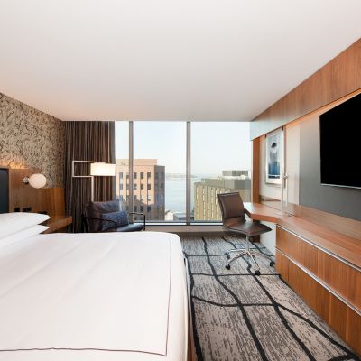 A modern hotel room with a large bed, a desk, a chair, a flat-screen TV, and a large window with a city view.