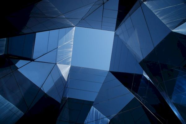 The image shows a geometric architectural structure made of reflective glass panels, forming a symmetric open space with the sky visible above.
