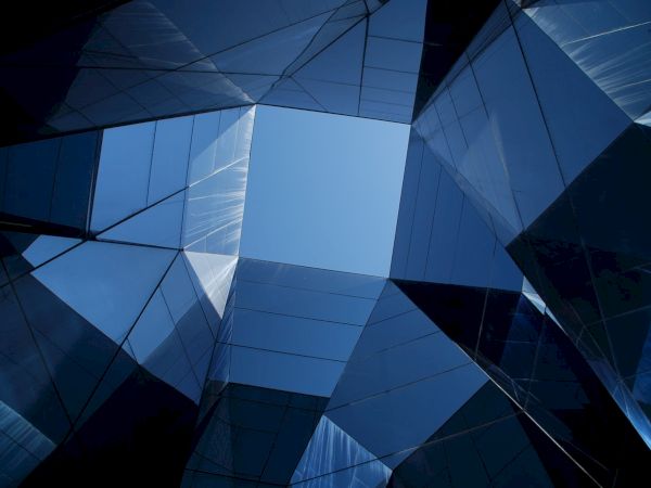 The image shows a geometric architectural structure made of reflective glass panels, forming a symmetric open space with the sky visible above.
