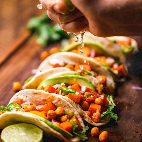 A hand is squeezing lime juice onto a row of tacos filled with vegetables and chickpeas, garnished with lime slices and fresh herbs, on a wooden board.