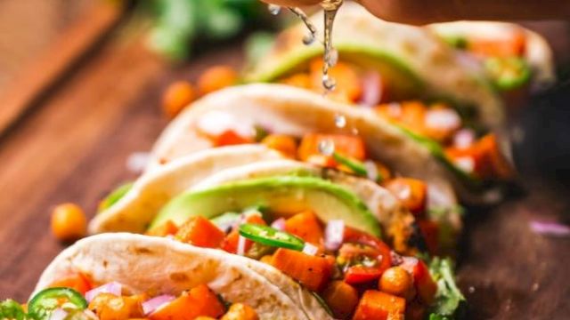 A hand is squeezing lime juice onto a row of tacos filled with vegetables and chickpeas, garnished with lime slices and fresh herbs, on a wooden board.