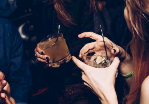 A group of people in a dimly lit setting holding drinks, engaged in conversation and laughter, with their faces partially visible.