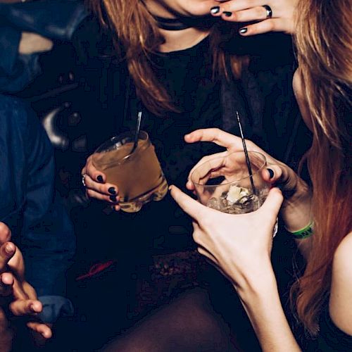 A group of people in a dimly lit setting holding drinks, engaged in conversation and laughter, with their faces partially visible.