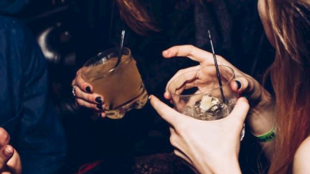 A group of people in a dimly lit setting holding drinks, engaged in conversation and laughter, with their faces partially visible.