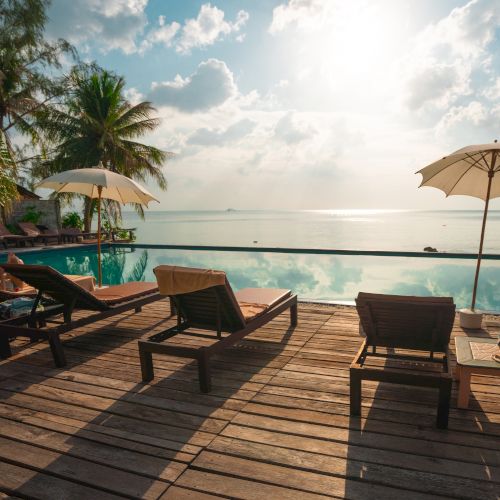 A serene beachside scene with lounge chairs and umbrellas on a wooden deck, overlooking a tranquil sea under a partly cloudy sky.