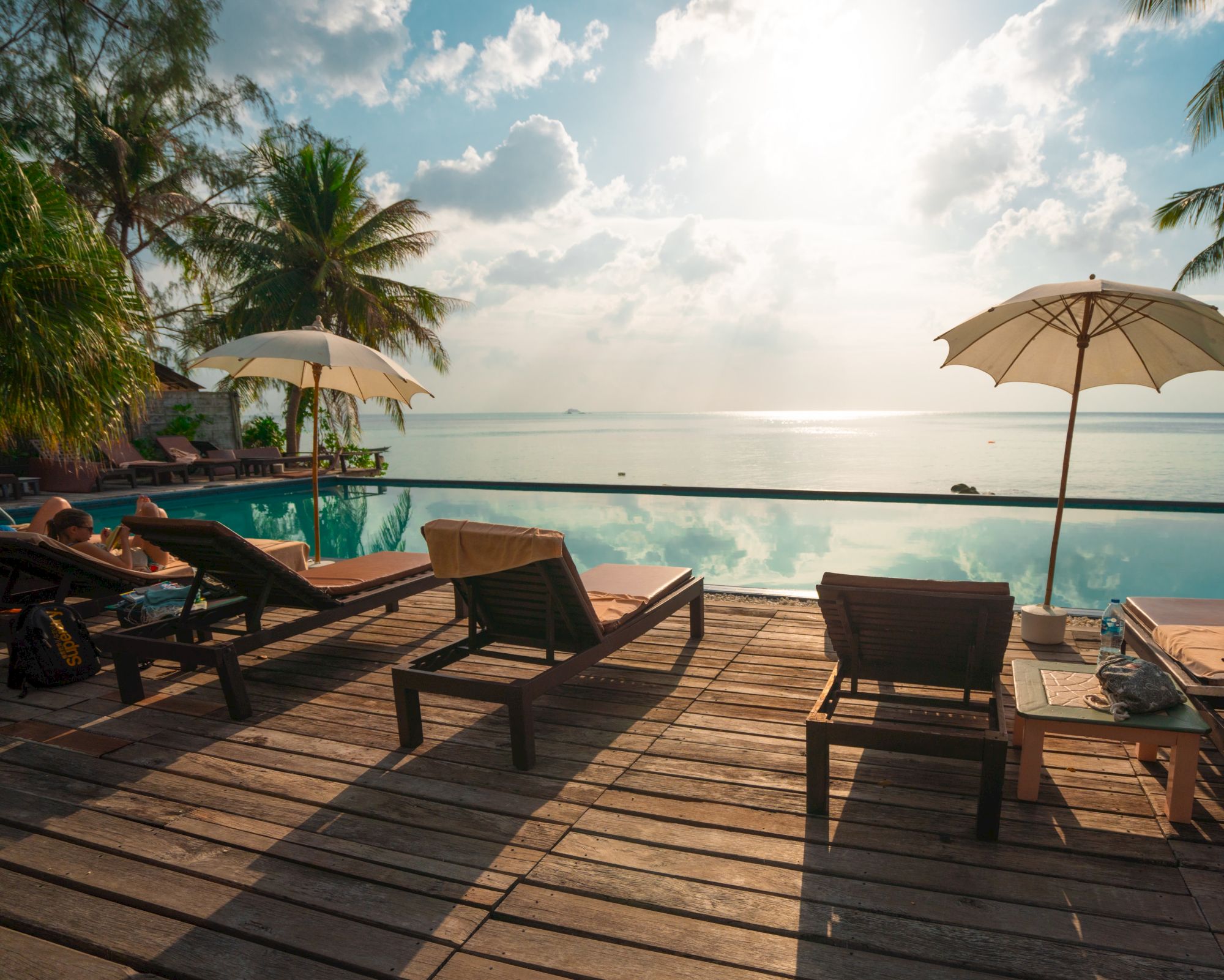 A serene beach scene with lounge chairs on a wooden deck, parasols, a glistening pool, palm trees, and a stunning sea view under a bright sky.