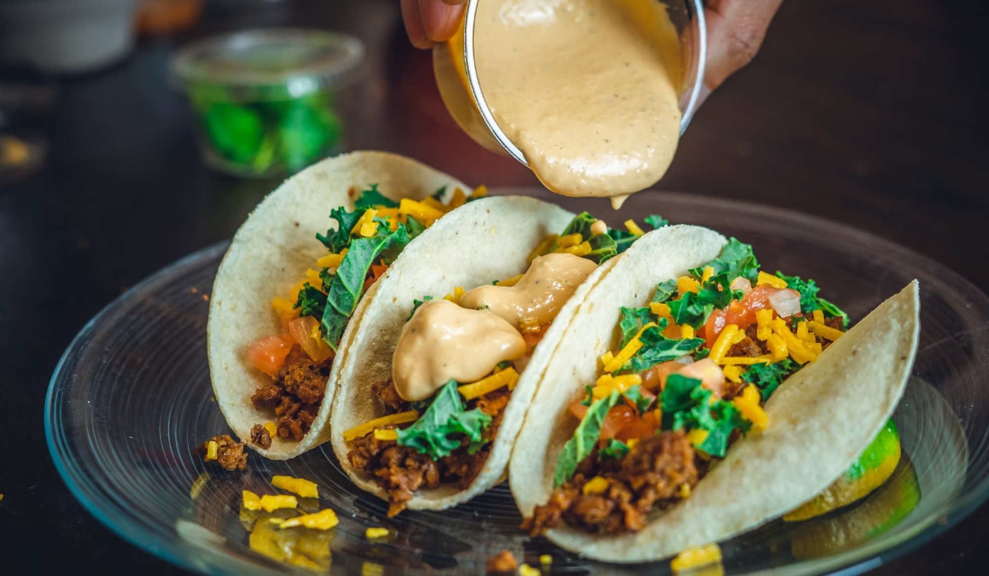 A hand pouring sauce onto three soft shell tacos filled with ground meat, cheese, tomatoes, and greens on a glass plate.