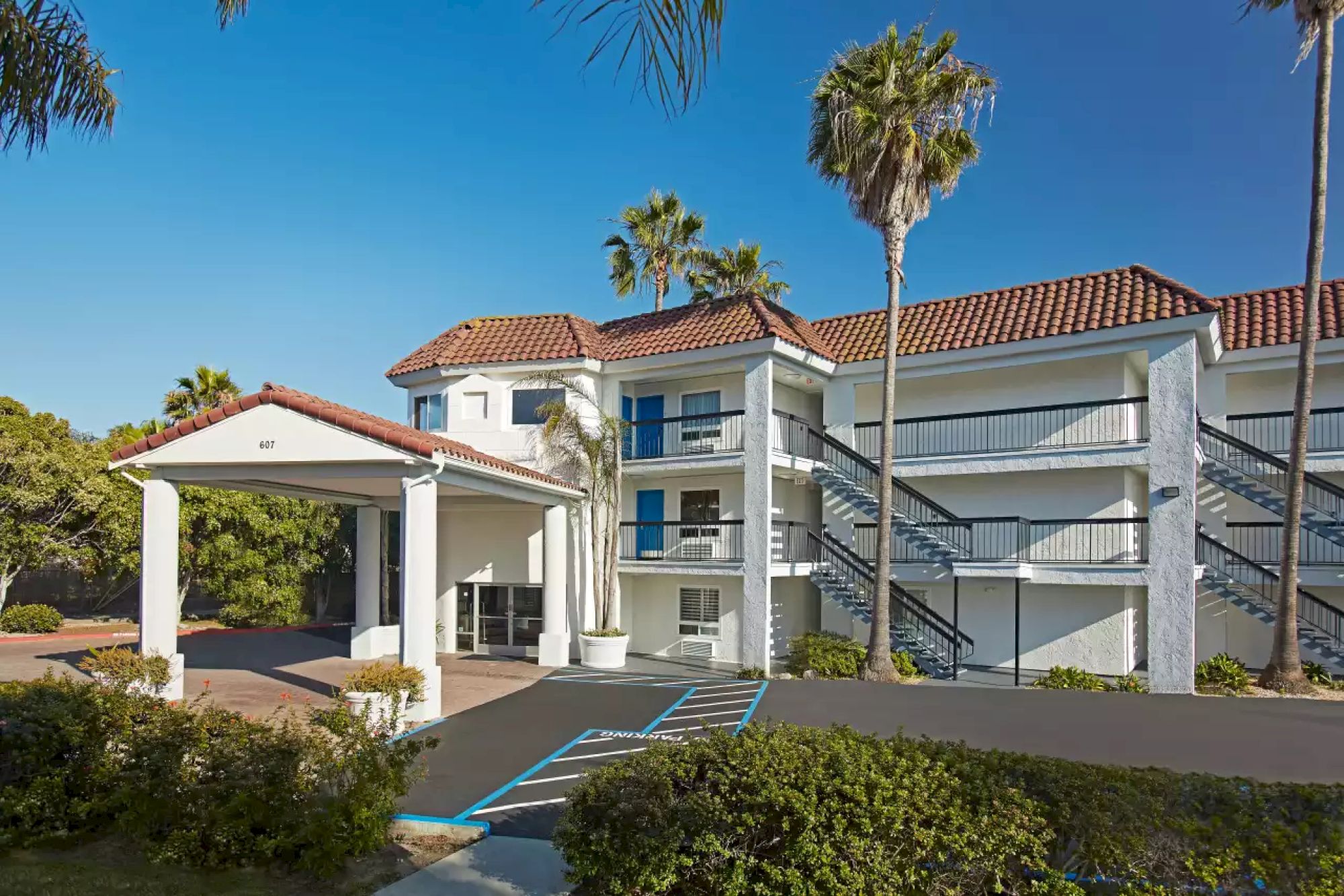 A three-story building with a red-tile roof, external staircases, and a covered entrance, surrounded by palm trees and greenery on a sunny day.