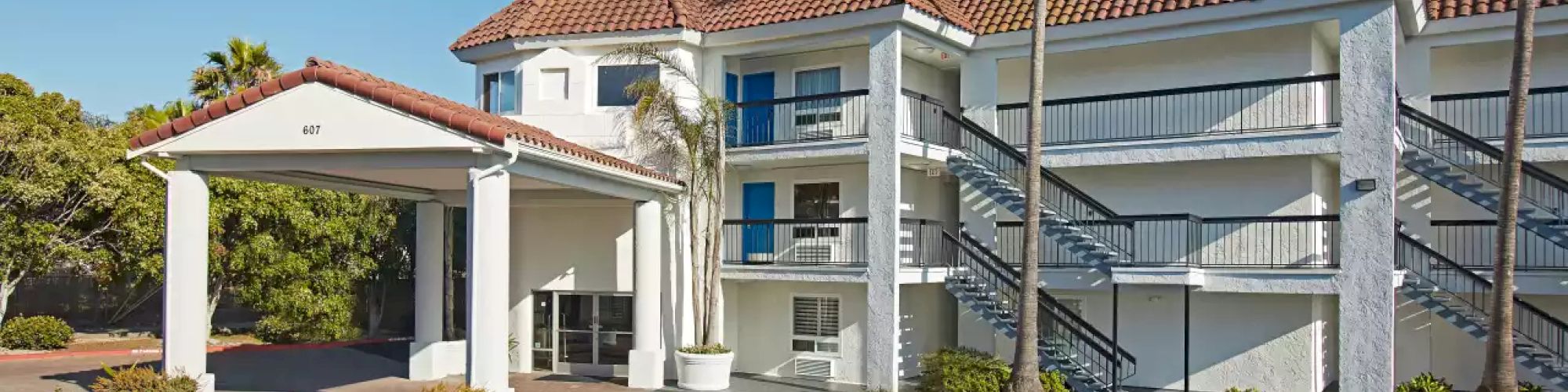 The image shows a two-story building with a red-tiled roof, exterior staircases, and palm trees around. The setting appears sunny.