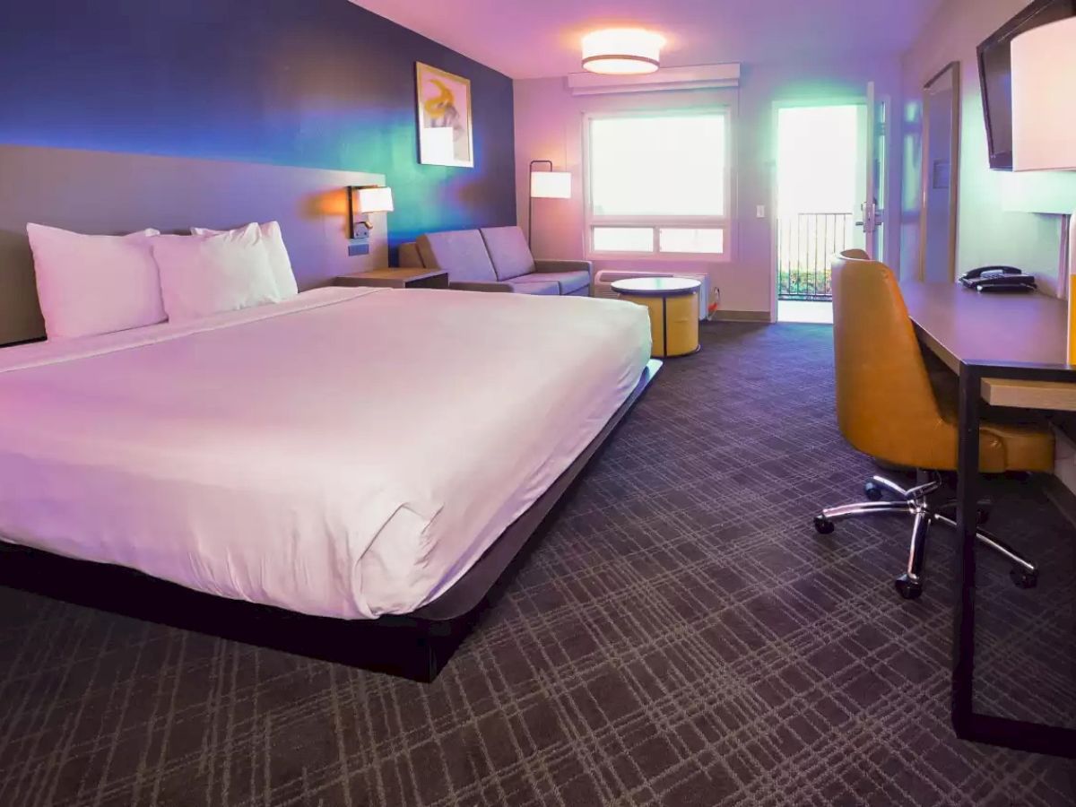 This image shows a modern hotel room with a large bed, desk, chair, sofa, table, and a window with natural light.