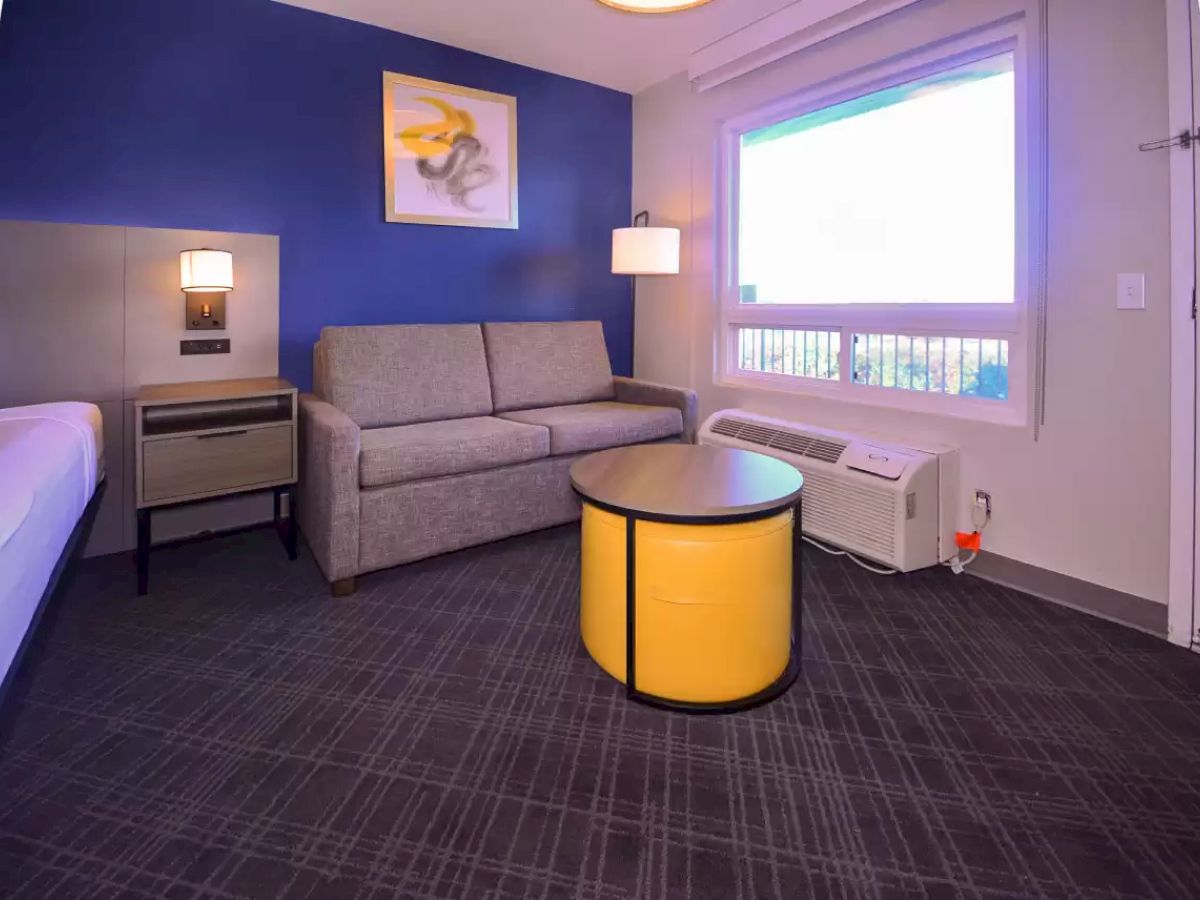 A modern room with a sofa, large round table, bed, nightstand, wall art, air conditioner, and window with a view, highlighted by blue and grey tones.