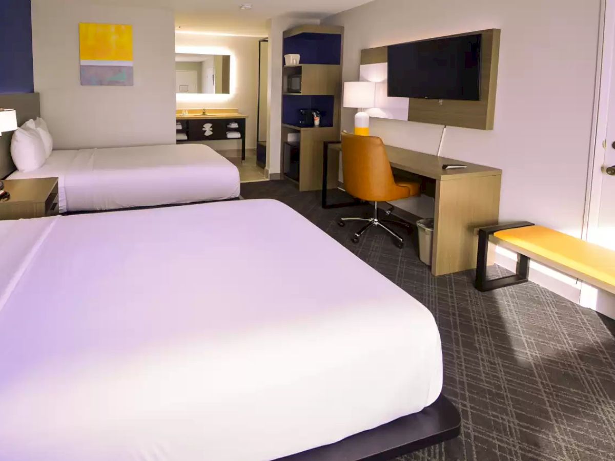 This image shows a hotel room with two beds, a desk with an orange chair, a wall-mounted TV, a bench, and modern decor.