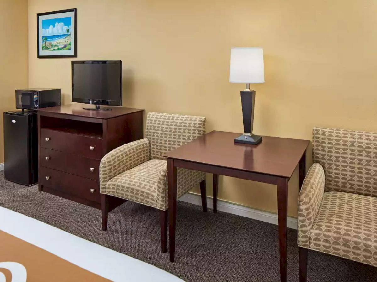 The image shows a hotel room with beige walls, a TV on a dresser, a microwave, two chairs, a table, a lamp, and framed artwork.