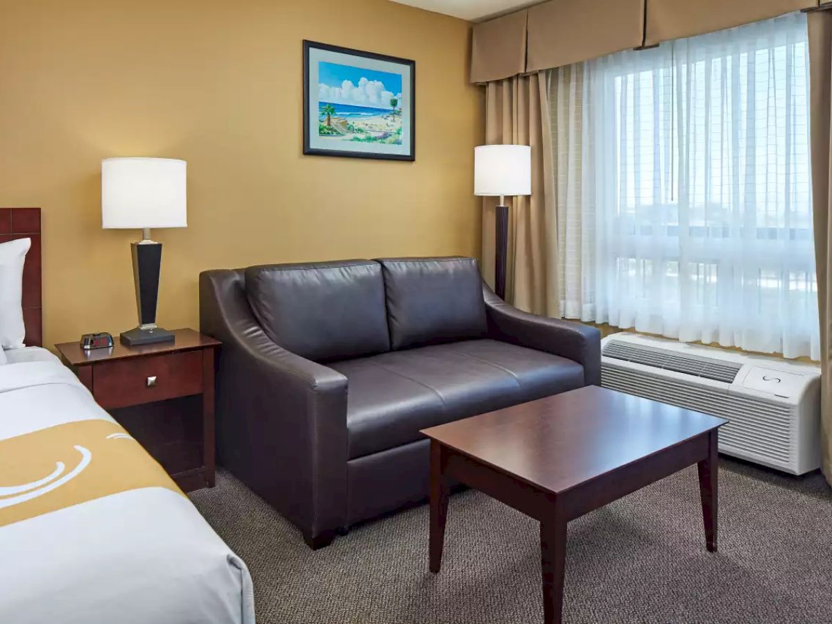 The image shows a hotel room with a bed, nightstand, lamps, a sofa, coffee table, window with curtains, air conditioning unit, and framed artwork.