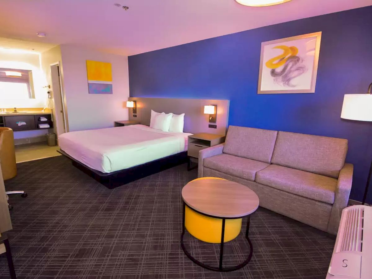 A modern hotel room with a bed, a sofa, a round table, wall art, and a workspace near the entrance to the bathroom, featuring warm lighting.