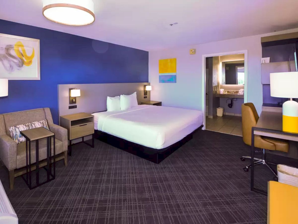 This image shows a modern hotel room with a bed, armchair, desk, and a bathroom. Wall art and lamps decorate the space, creating a cozy atmosphere.