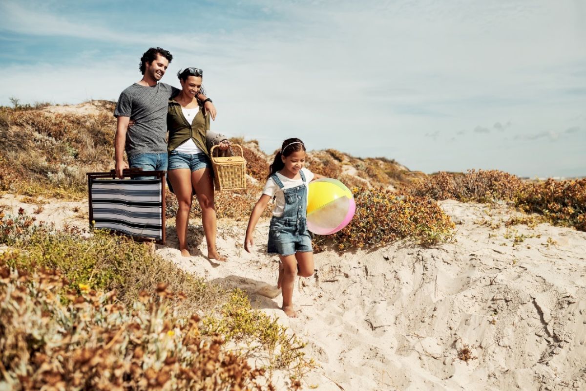 A family walks down a sandy path towards the beach, carrying a picnic basket, bag, and a colorful inflatable beach ball.