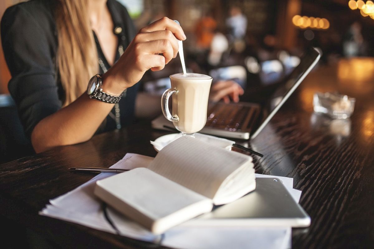 A person is sitting at a table with a laptop, stirring a drink in a glass cup while notebooks, a pen, and a cellphone rest on the table.