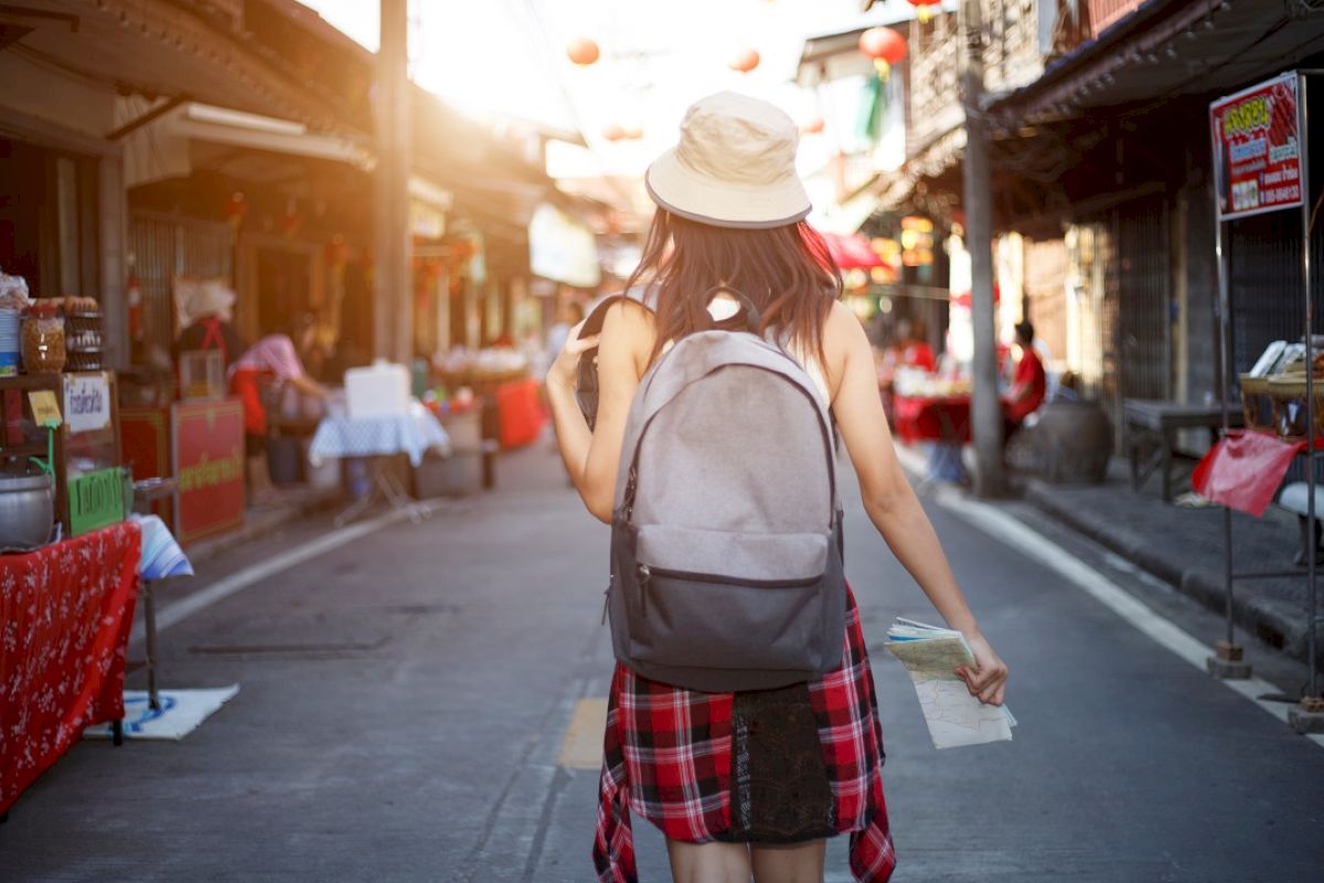 A person wearing a hat and backpack walks down a bustling street lined with market stalls, holding a map.