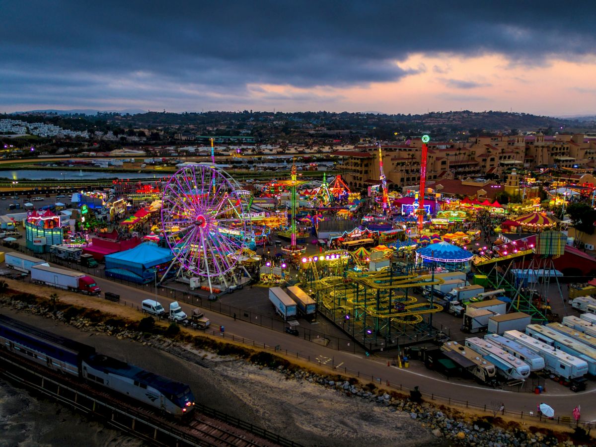 The image shows a brightly lit carnival at twilight with various rides, including a Ferris wheel, surrounded by trucks and trailers.