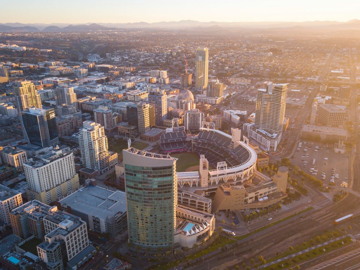 The image shows an aerial view of a cityscape at sunset, featuring a large stadium centrally located among high-rise buildings, with mountains in the distance.