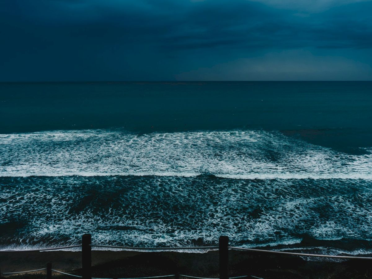 The image shows a dark, stormy ocean with waves crashing on the shore under a moody, cloudy sky, and a wooden fence in the foreground.