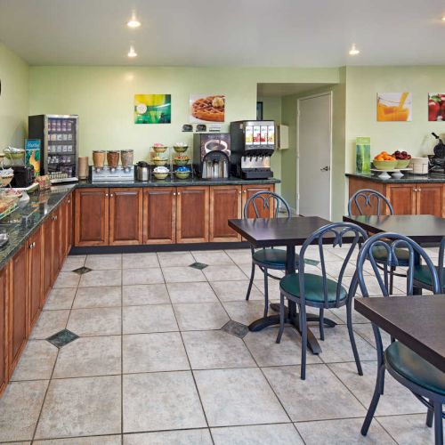 The image shows a breakfast area with food counters, various breakfast items, a coffee machine, chairs, tables, and colorful wall art.