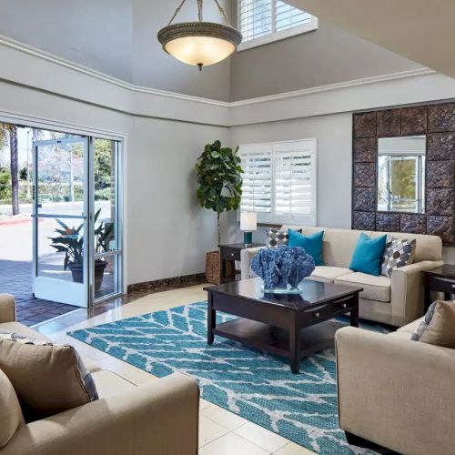 A modern living room with beige sofas, blue accents, a large rug, indoor plants, and a central coffee table, near a glass door to the outside.