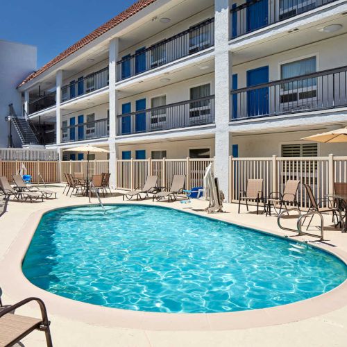 The image shows an outdoor pool area with lounge chairs, umbrellas, and a three-story building in the background, likely a hotel or motel.