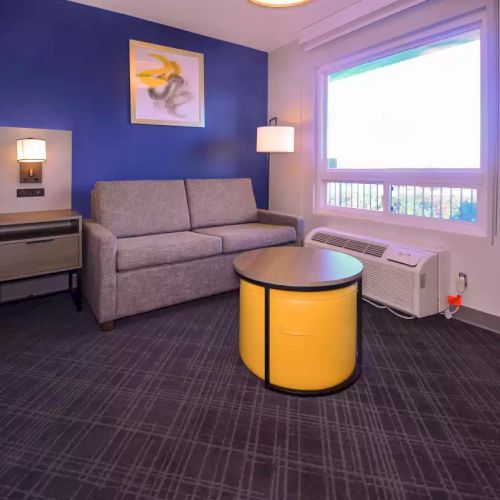 A hotel room features a bed, a gray sofa, a yellow round table, an air conditioner, a nightstand, a lamp, and a window with a view.