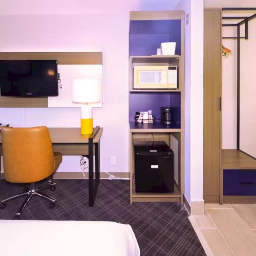 A hotel room with a desk, chair, TV, lamp, microwave, mini-fridge, coffee maker, and open closet area.