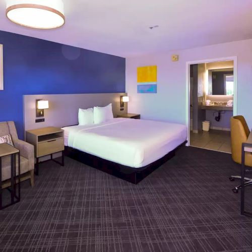 This image shows a modern hotel room with a king-sized bed, armchair, desk, and a bathroom. The decor features blue walls and abstract artwork.