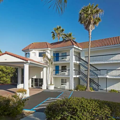 The image shows a two-story building with exterior stairs, palm trees, and landscaped surroundings, featuring a tiled roof and multiple balconies.