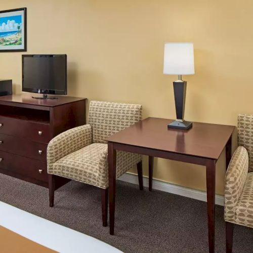 A cozy room with a bed, a table with a lamp, two chairs, a TV on a dresser, a mini fridge, and a wall picture.